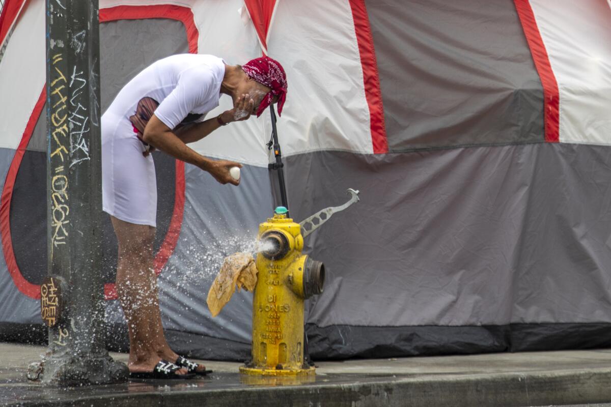 A woman wearing a red bandanna washes her face near a fire hydrant, with a gray tent in the background