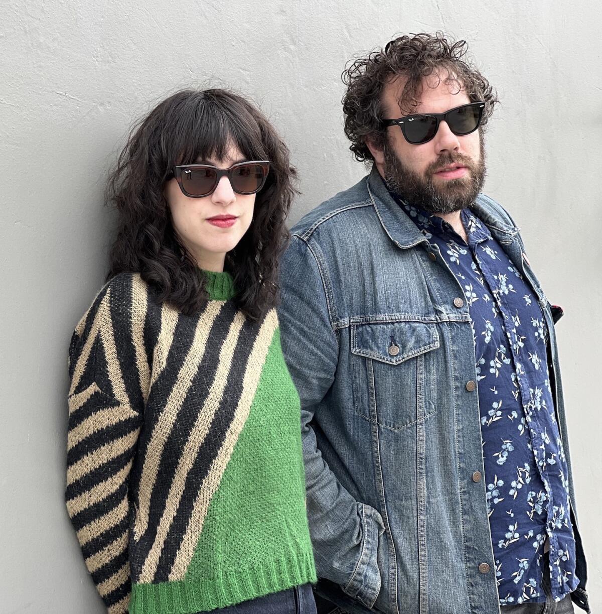 a woman, left, in a sweater and a man in a jean jacket and print button-down lean against a wall. both wear sunglasses 