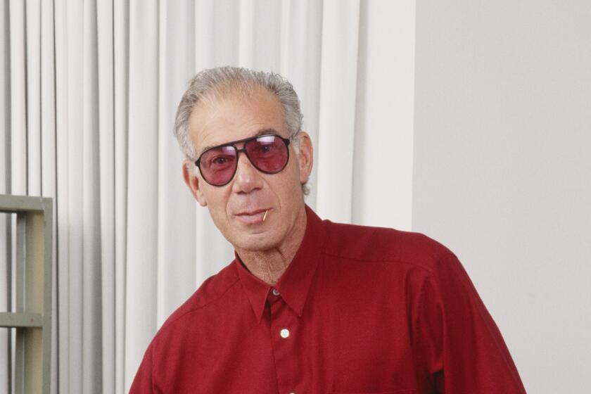 A man posing in gray sunglasses, gray pants and a red, button-up shirt