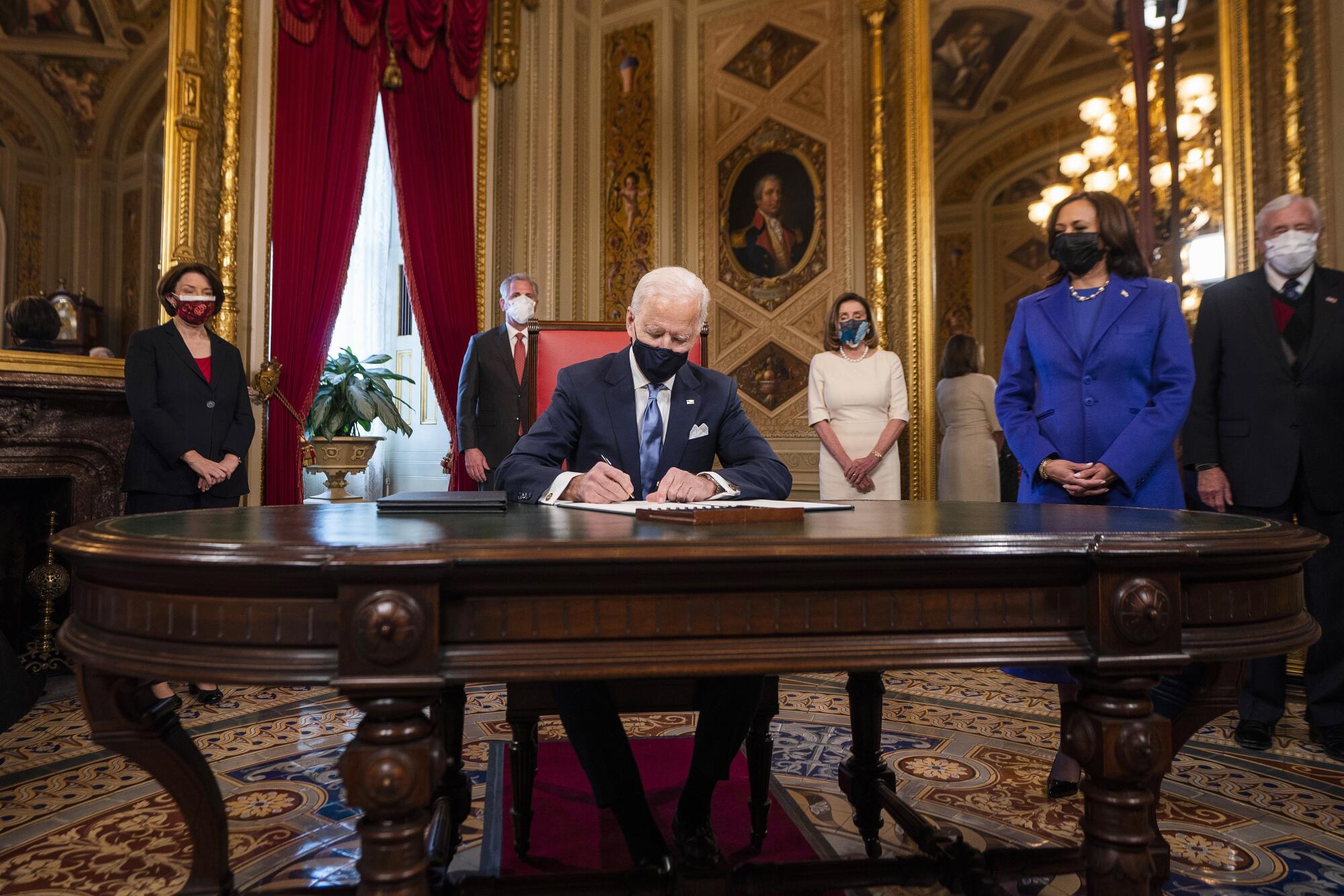 Biden, at an ornate table, signs documents. Among those standing, socially distanced, is Kamala Harris.