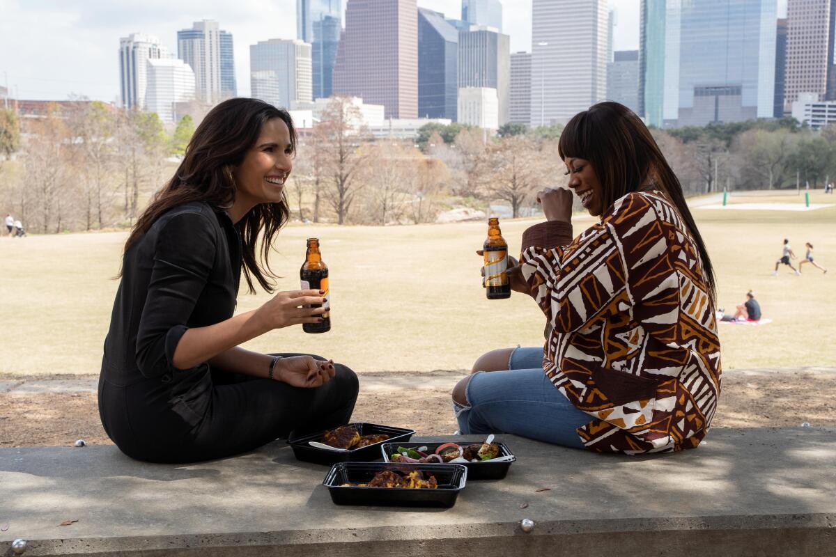 Padma Lakshmi and Yvonne Orji laughing and drinking beer outdoors, the Houston skyline in the background