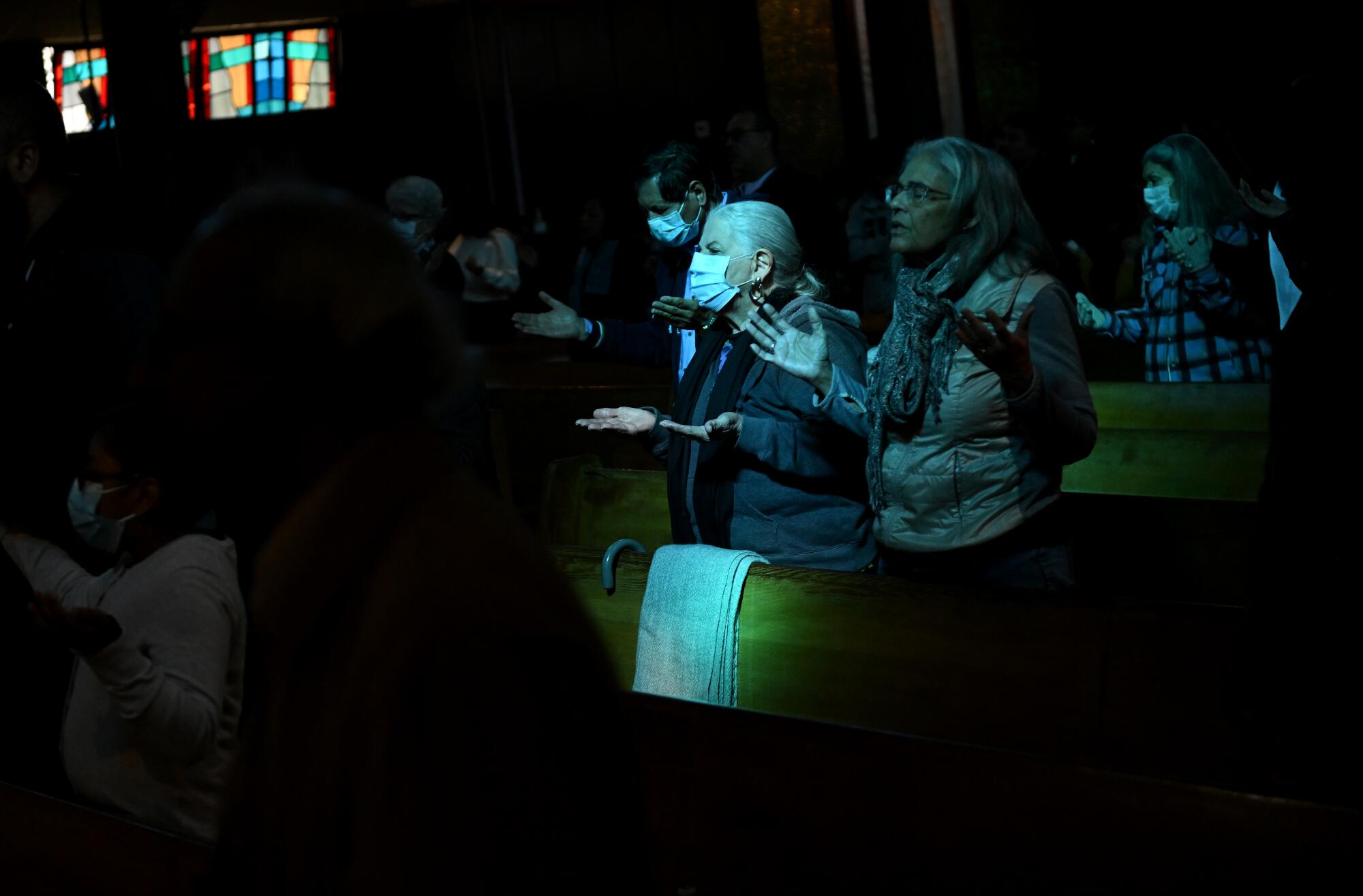 Standing in darkened pews, lit by blu-tinted light coming in through a stained-glass window, people pray during a mass