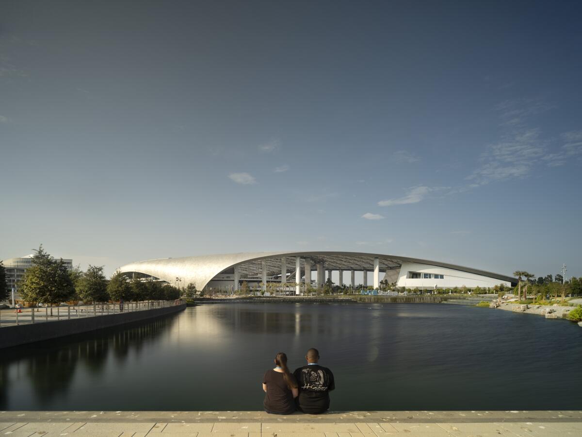 A couple sits on an overlook with views of a lake and SoFi Stadium's curving roof gleaming in the sun