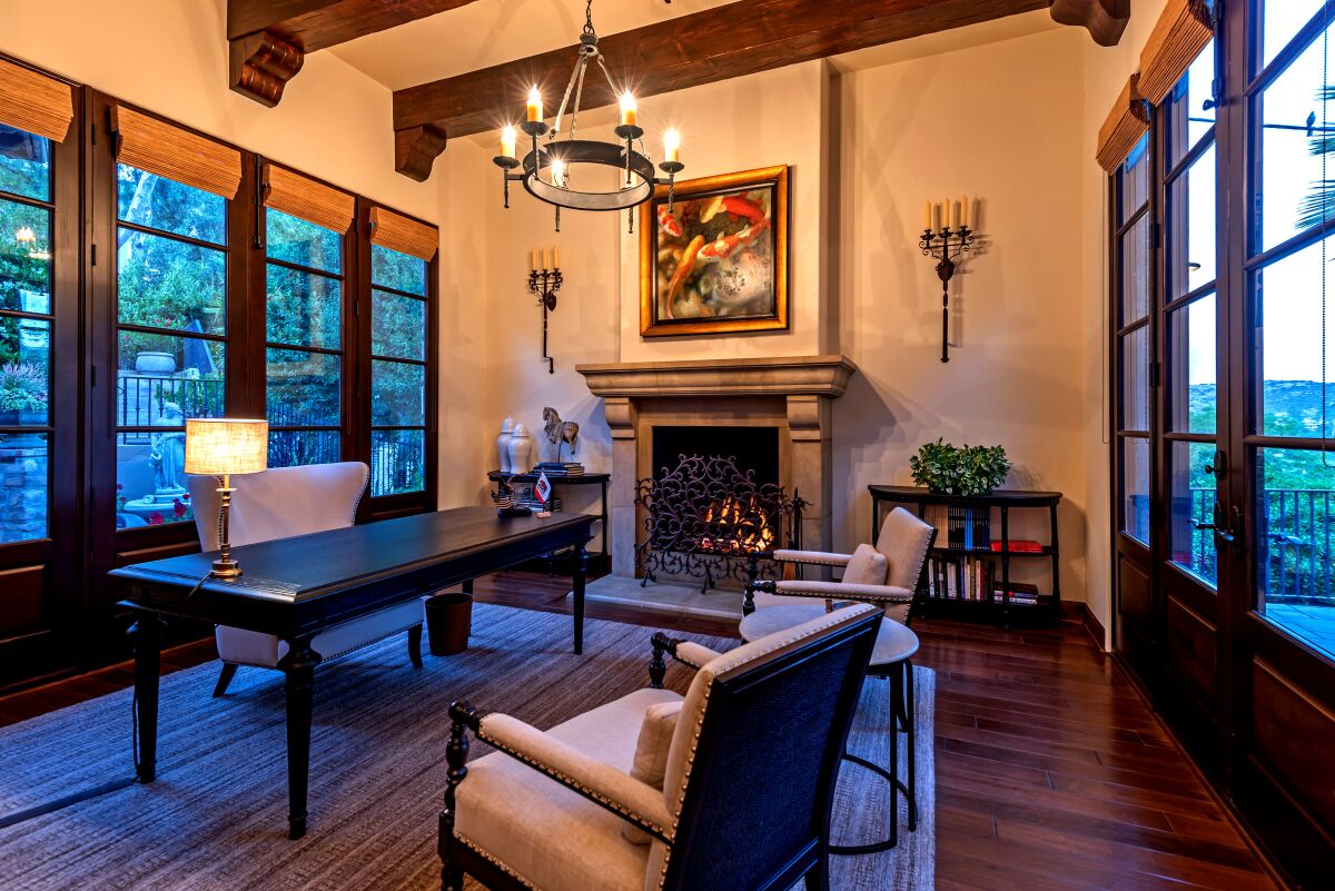 An office in the Mediterranean villa-style house has dark wood beams, a fireplace, walls of windows.