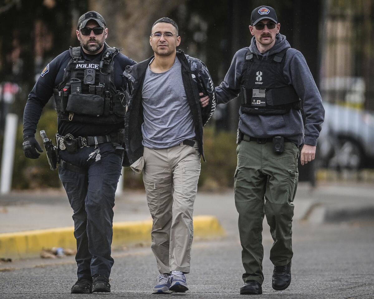 A man with dark hair and glasses is escorted by two men with dark vests, one of which says Police. 