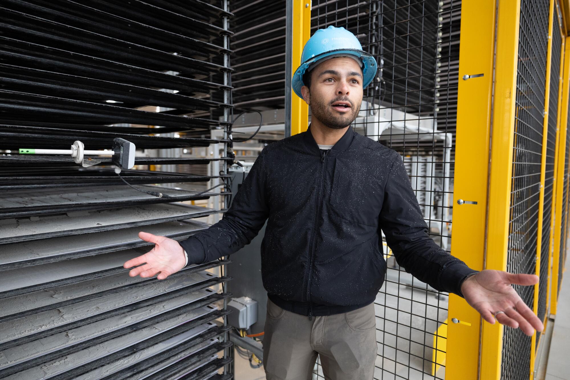 A man in a black jacket and blue hard hat stands beside a bank of trays 