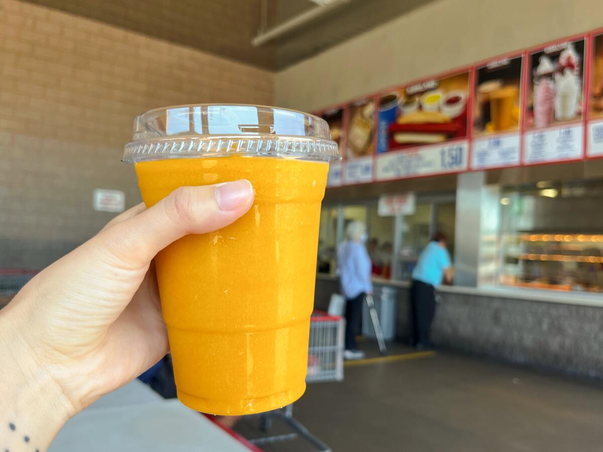 A hand holds up a clear plastic cup filled with orange mango smoothie