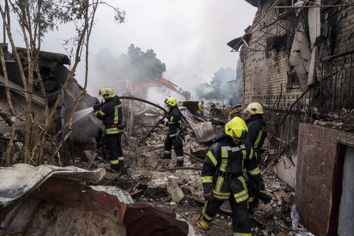 Workers in protective gear spray water on smoldering rubble.