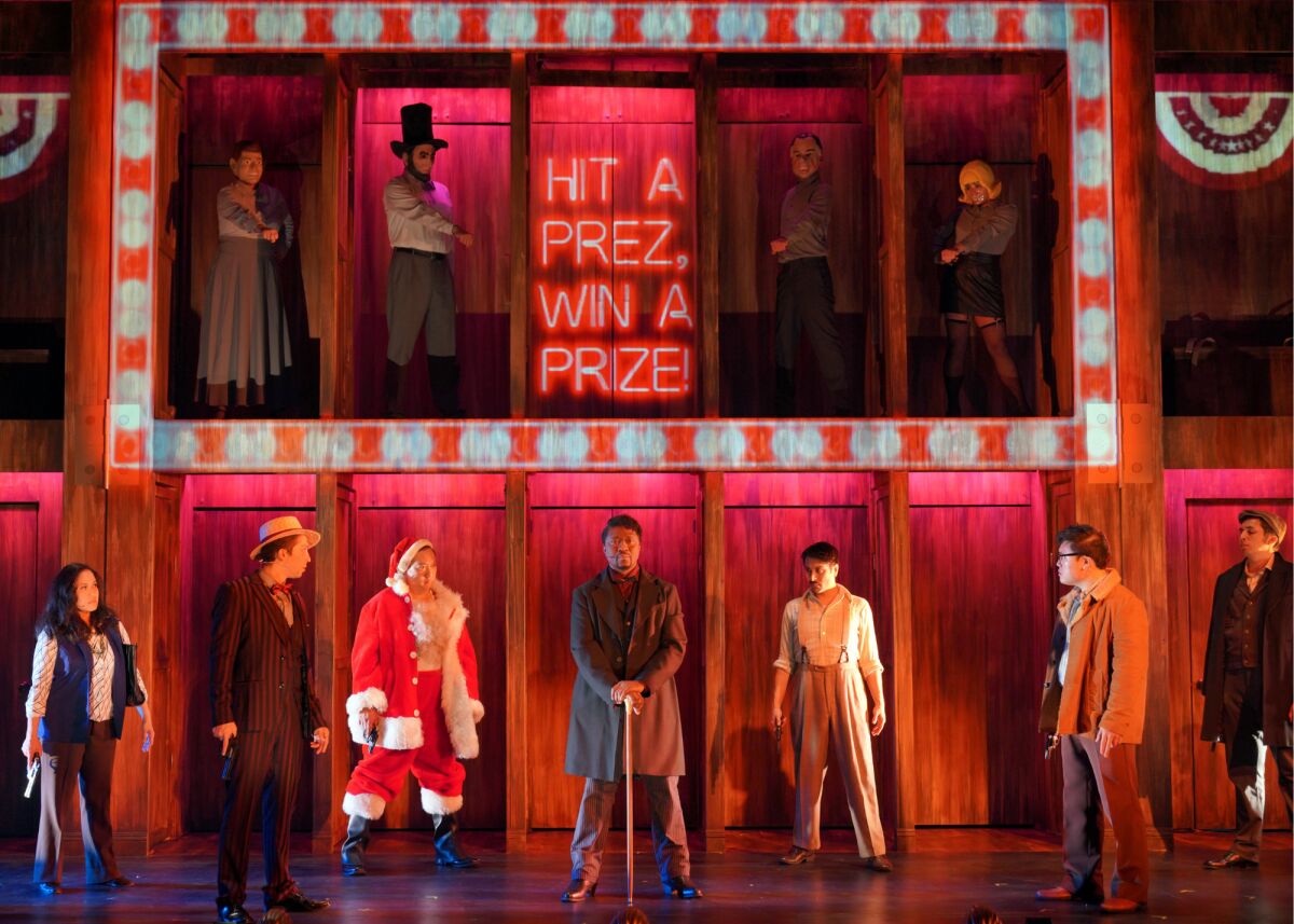 A group of people, including one in a Santa suit, stand in front of a set onstage with neon words "Hit a Prez, Win a Prize"