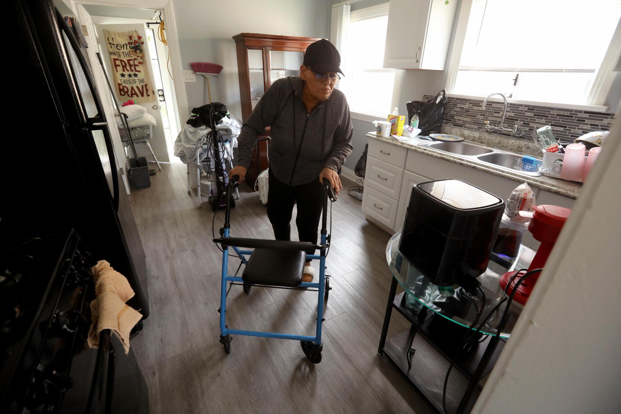 A woman uses a walker to make her way through the kitchen in her home.