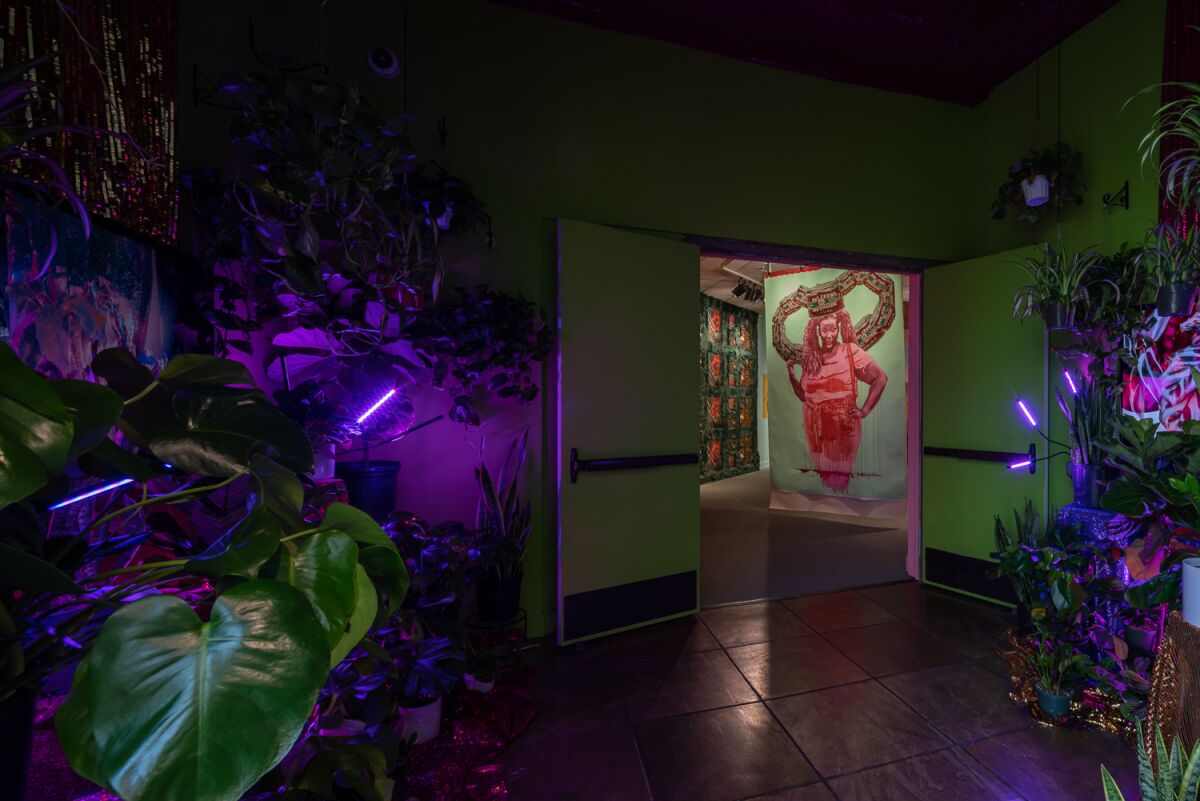 Plants and purple lights frame a doorway with the figure of a Black woman with a crown-like structure on her head.