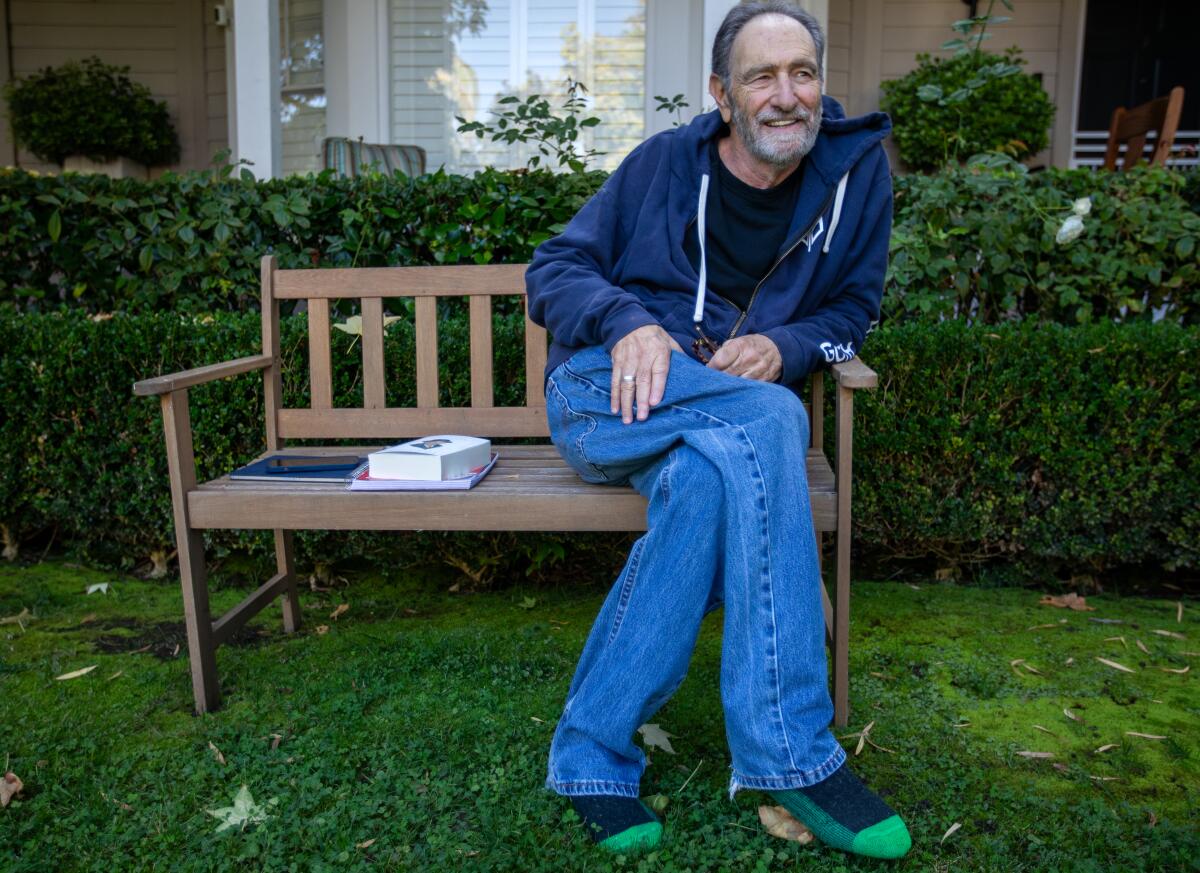 A man in jeans and a hoodie sits on a bench with his legs crossed.