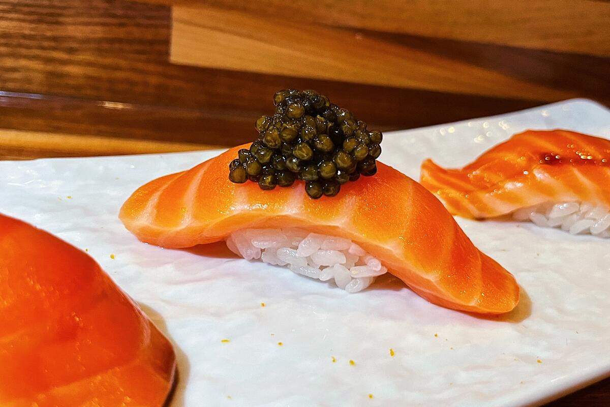 Three salmon nigiri on a white plate. In the center, the Ora King belly cut features a mound of Astrea caviar atop it.