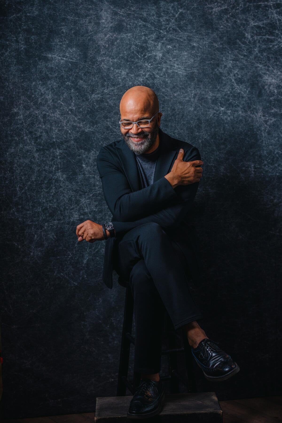 Jeffrey Wright crosses his arms, sitting on a stool and looking down in a portrait.