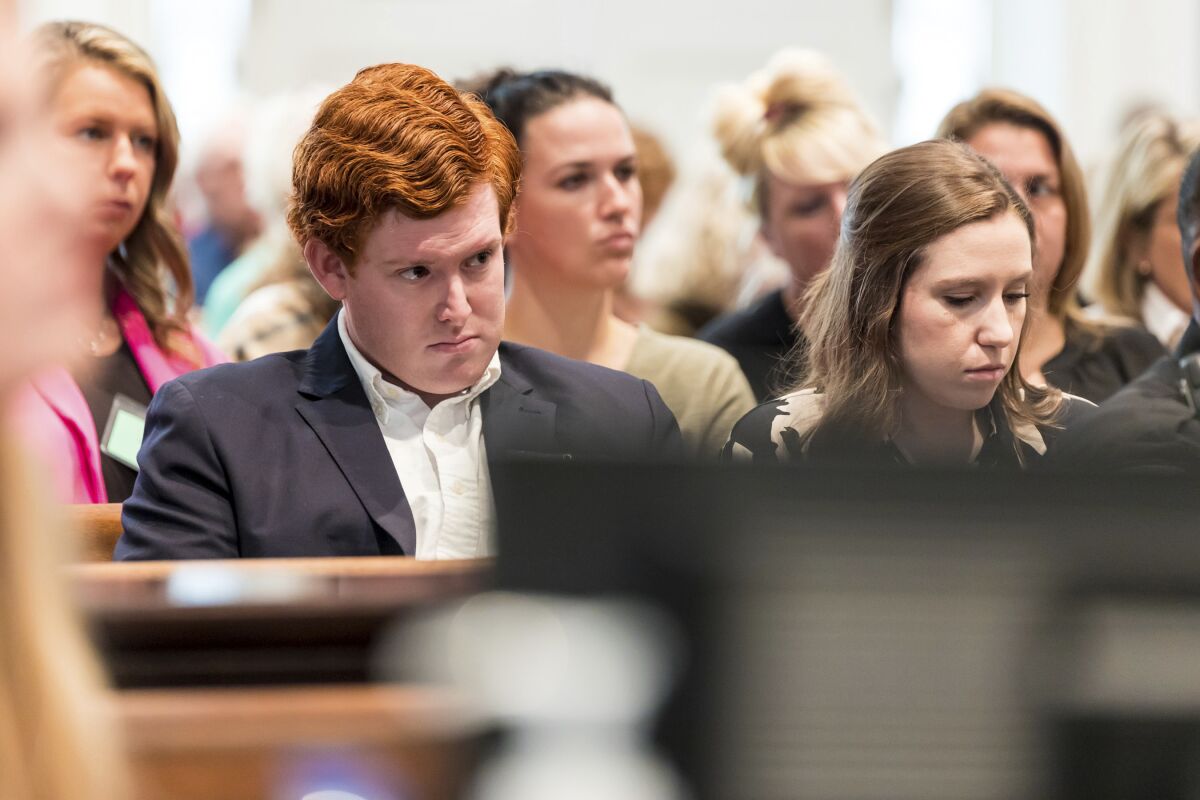 A young man with red hair sits on a bench in a courtroom next to a young woman with dark hair