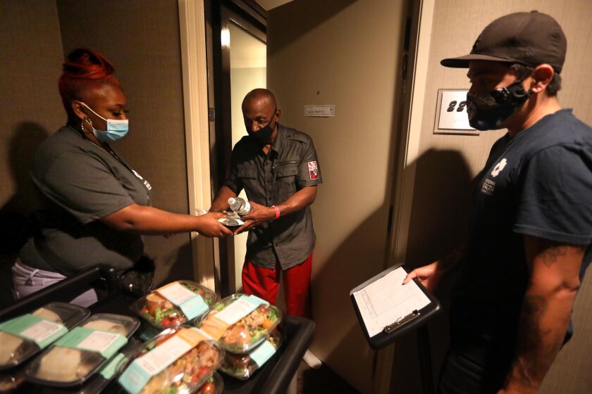 LOS ANGELES, CA - JULY 21, 2020 - - Guest services associate Mia Rogers, 23, left, gives Project Roomkey guest Stanley Monk, 58, his lunch under the watchful eye of LVN Richard F. at a Project Roomkey hotel in Los Angeles on July 22, 2020. Rogers, a client of Chrysalis, lost her job due to the coronavirus pandemic. She was hired to work at a Project Roomkey hotel. About 100 clients of Chrysalis, a downtown agency that gets work for homeless people who lost their jobs because of the virus shutdowns. They've been rehired by Project Roomkey to work in hotels. (Genaro Molina / Los Angeles Times)