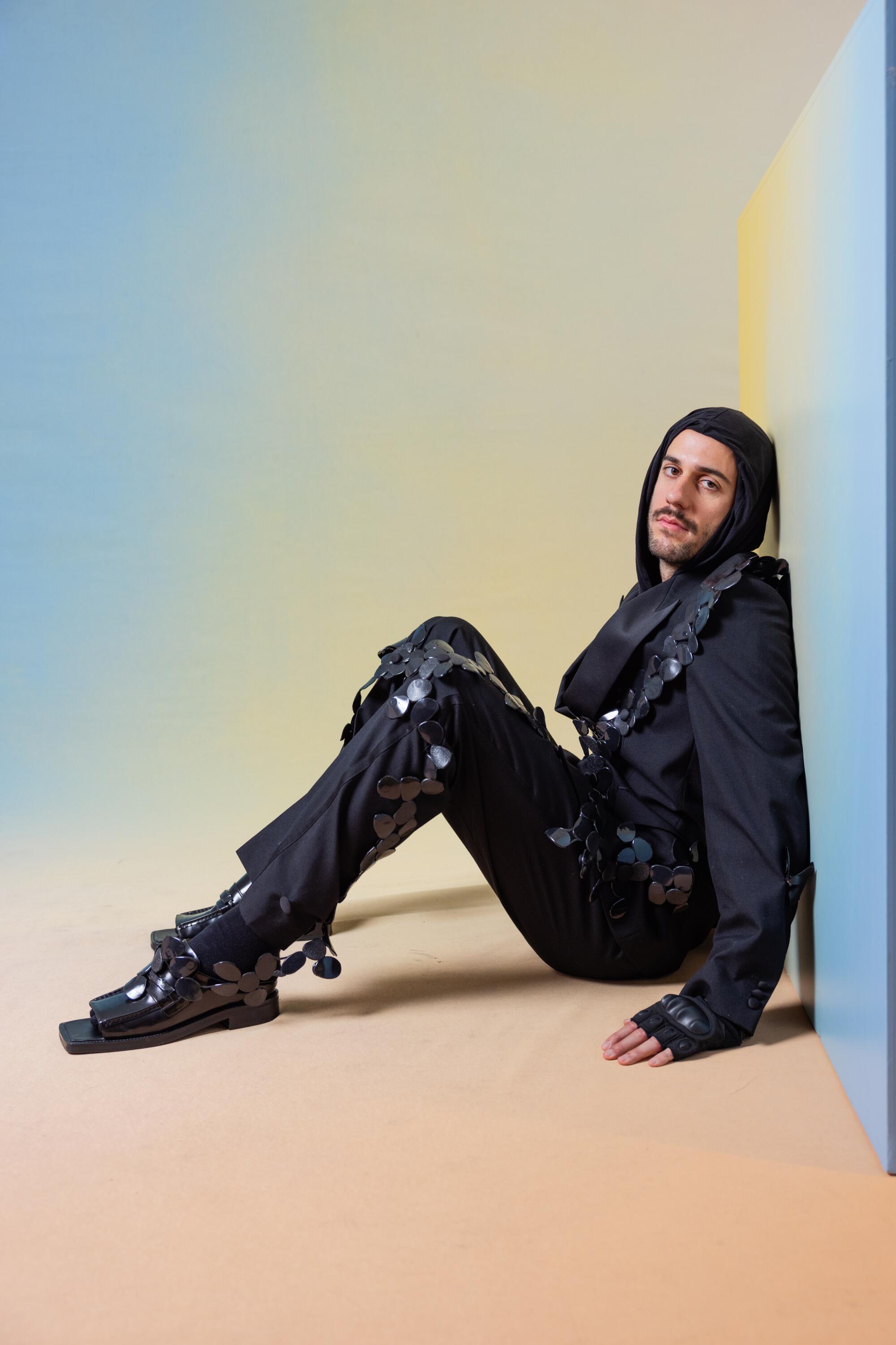 Aury Alby wears a dark outfit and hood while sitting on the floor and leaning against a wall.