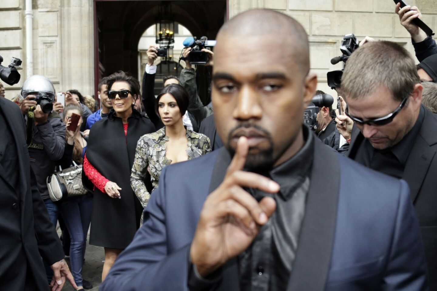 Kanye West appears to drop a hint to the crowd as he, Kim Kardashian and Kris Jenner, left, leave the George V hotel in Paris with baby North West on their way to Valentino's castle for brunch.