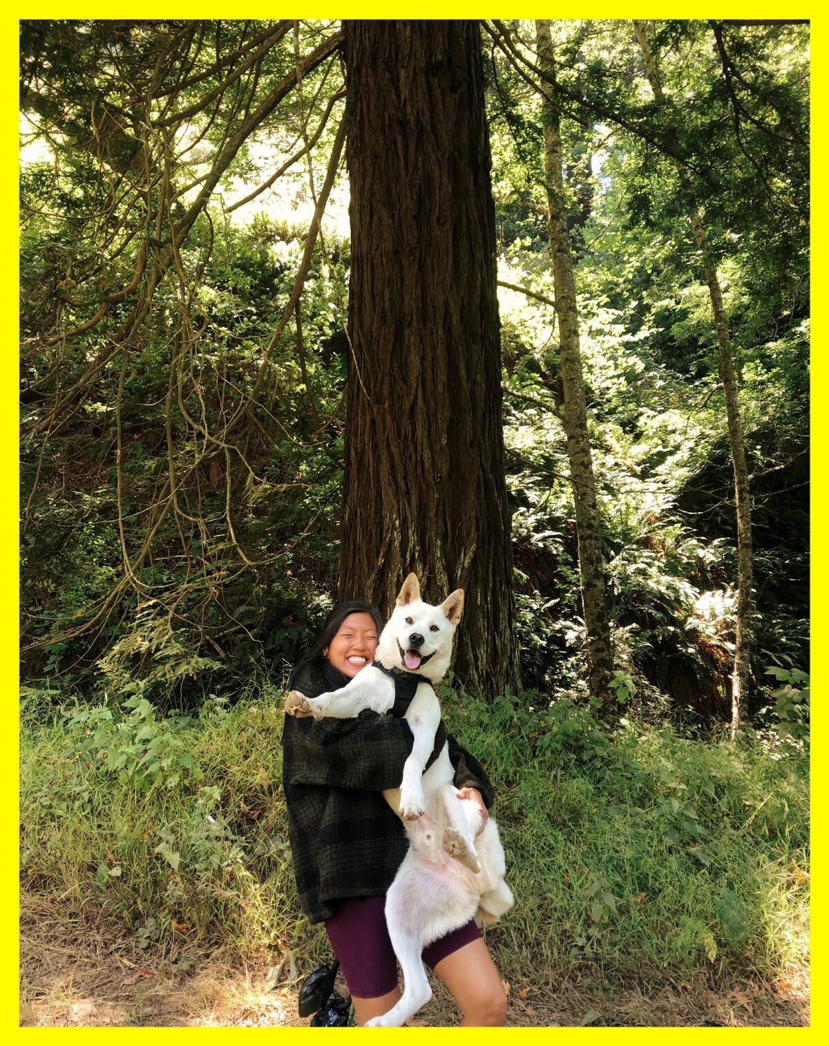 A woman holds her white dog in the outdoors. Both are smiling.