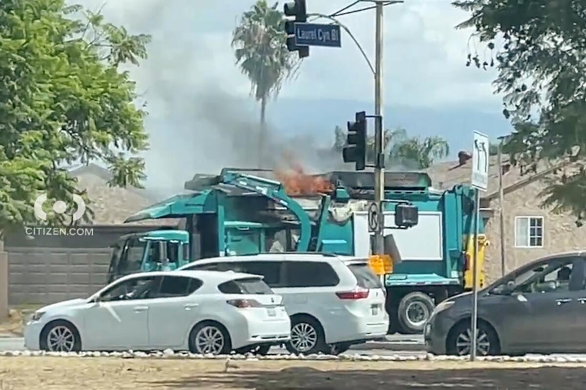 A sanitation truck on fire in a neighborhood nears cars and trees