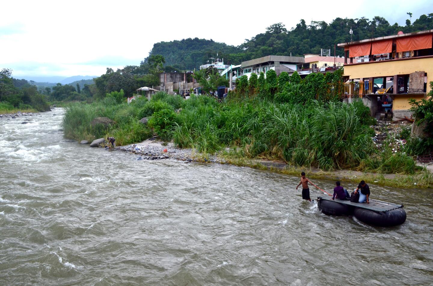 A boatman hauls an inner-tube raft and two men on the Suchiate River near Talisman, Mexico. The riverbank with the buildings is the Guatemalan side; the official border crossing is on a bridge just a few yards away.