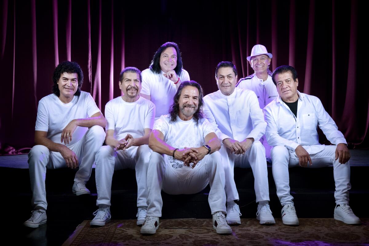 Seven men dressed in all white sit on the edge of a stage and smile