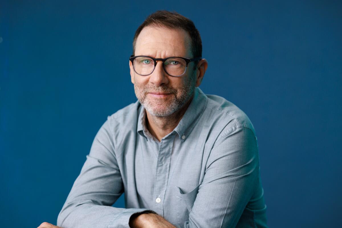 A bearded man with glasses, in a blue long-sleeved shirt, crosses his arms on a table.