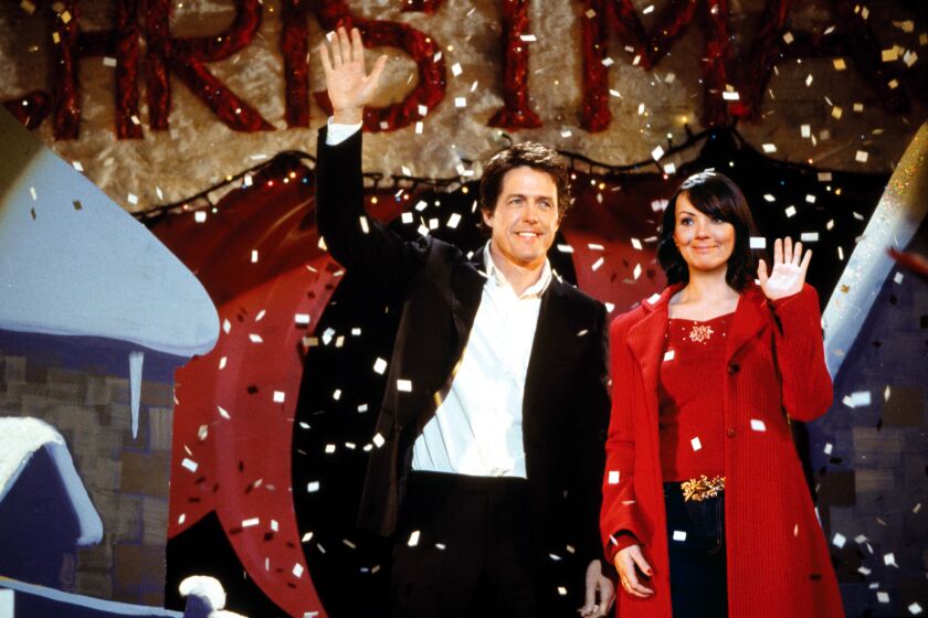 A man in a black suit and a woman in a red coat smiling and waving as fake snow falls around them