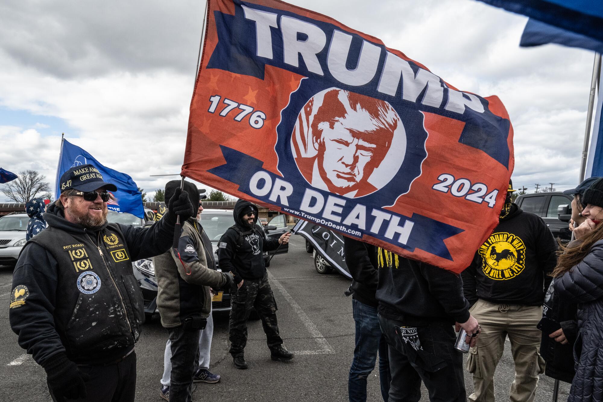People in a parking lot, some with flags, one reading "Trump or death" with 1776 and 2024 on either side of his image.