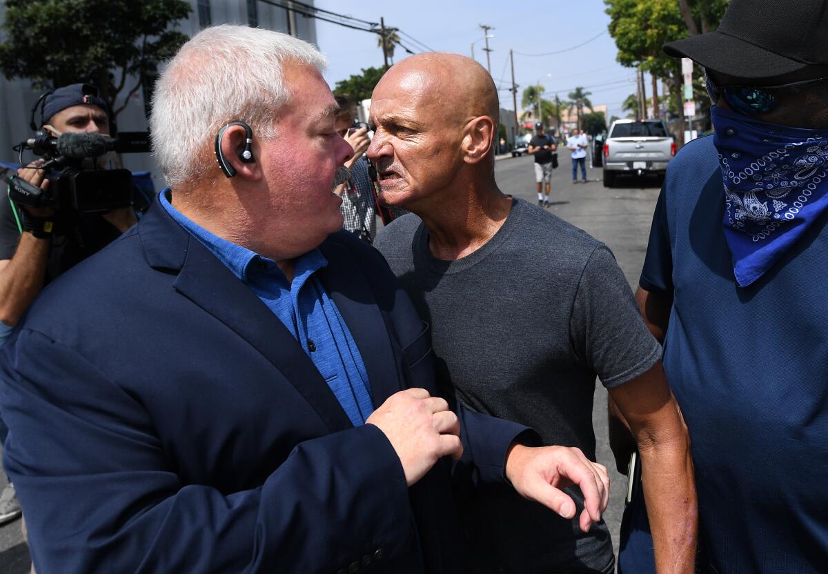 A man in an earpiece turns toward a bald, sneering man who is in his face.