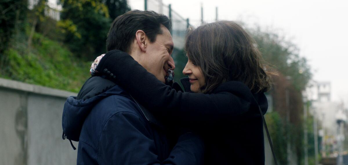 Standing outside, a man and woman in winter jackets smile as they embrace.