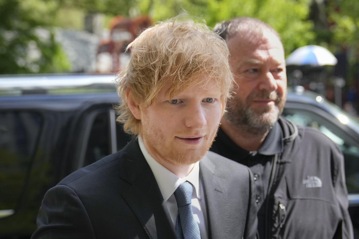 Ed Sheeran with shaggy red hair walks into court in dark blue suit and light blue tie.