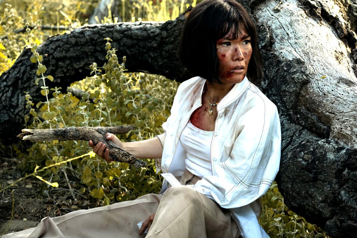 A woman with a bloody face in a white shirt and khakis sits near a tree with a stick in her hand.