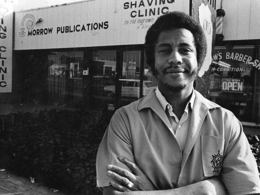 A black-and-white photo of a man posing in front of storefronts that say "Morrow Publications" and "Shaving Clinic"