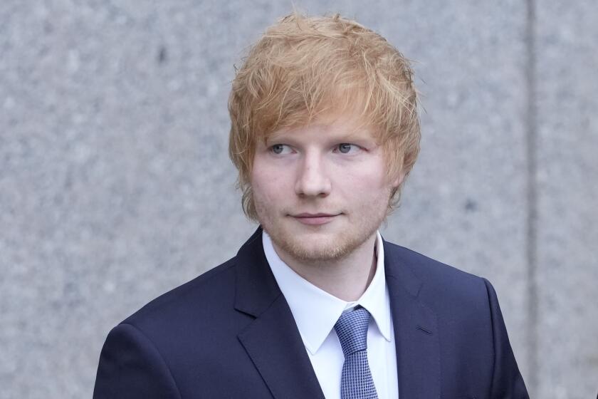 Ed Sheeran wearing a dark blue suit against a gray wall