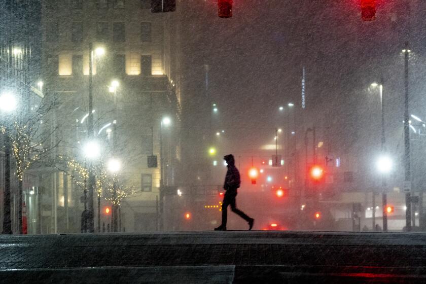 A silhouetted pedestrian walks across a street as snow falls and red street lights shine in the distance.