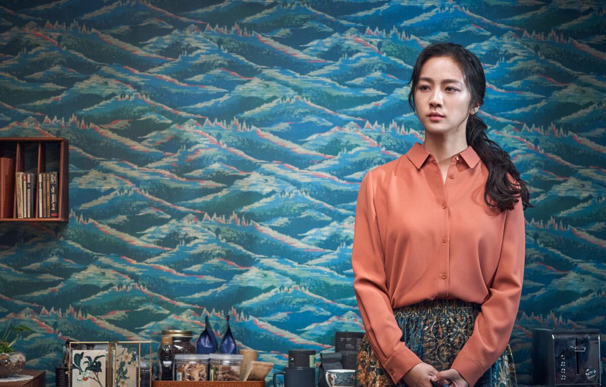 A woman in a blouse and skirt stands in front of a wallpapered wall 