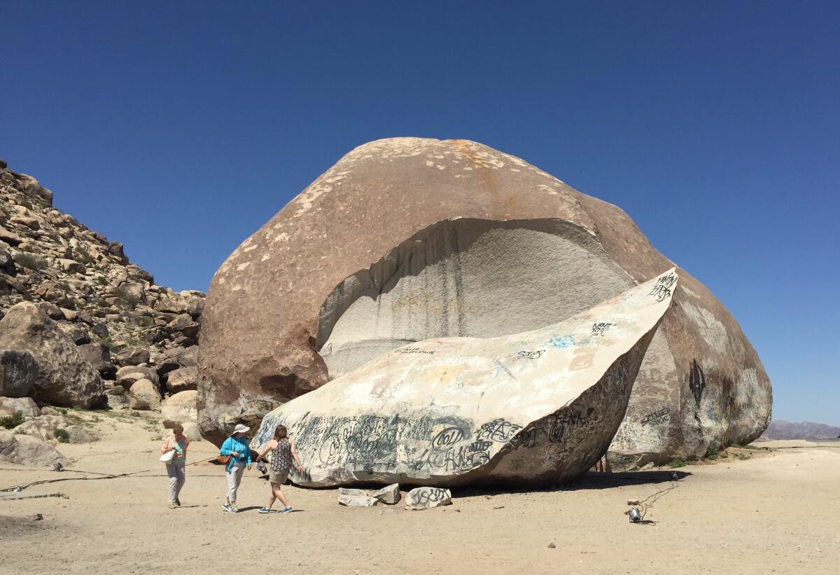 Three people stand in front of a massive boulder under a clear blue sky.