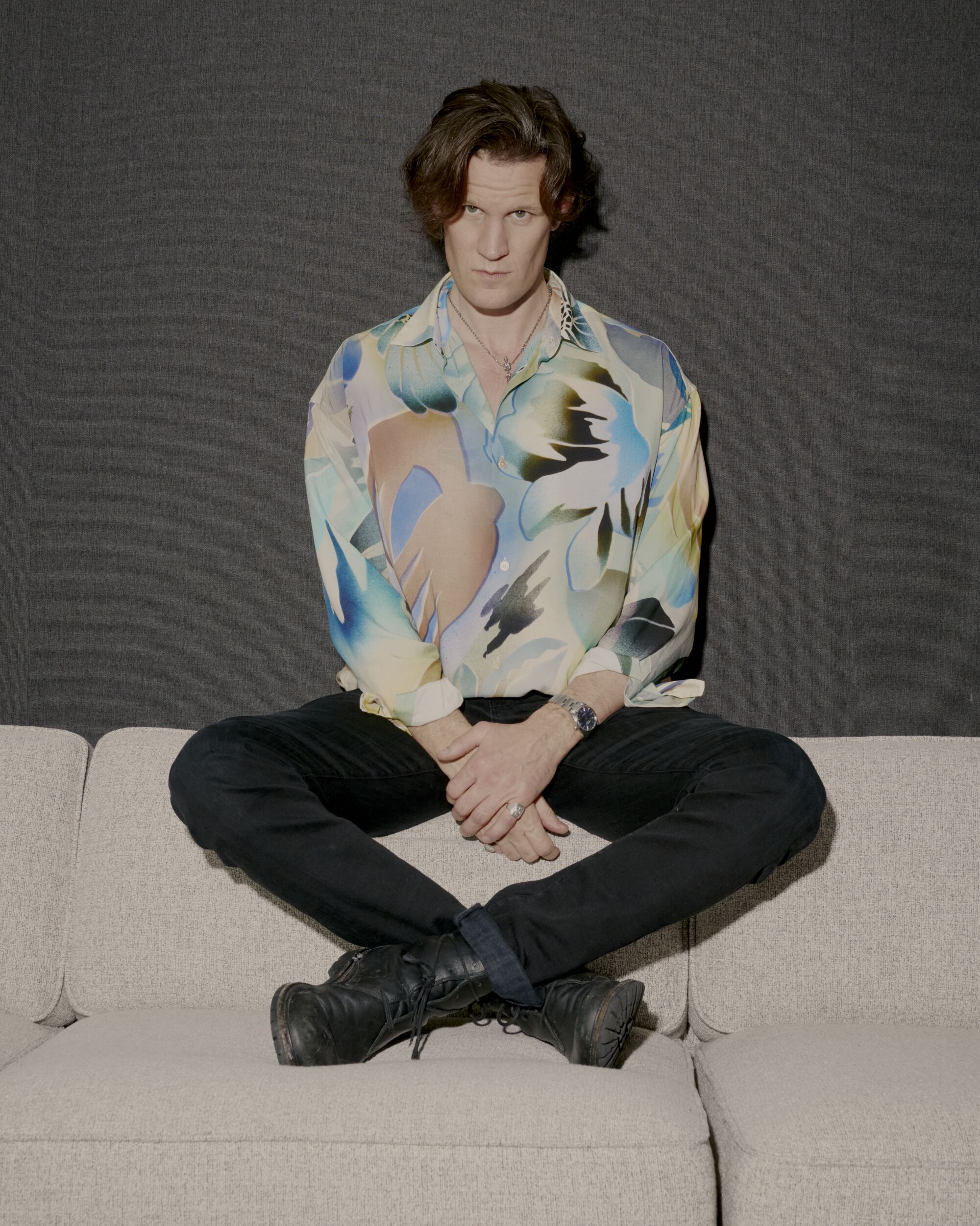 A man in black jeans and a colorful patterned shirt sits behind a sofa. 