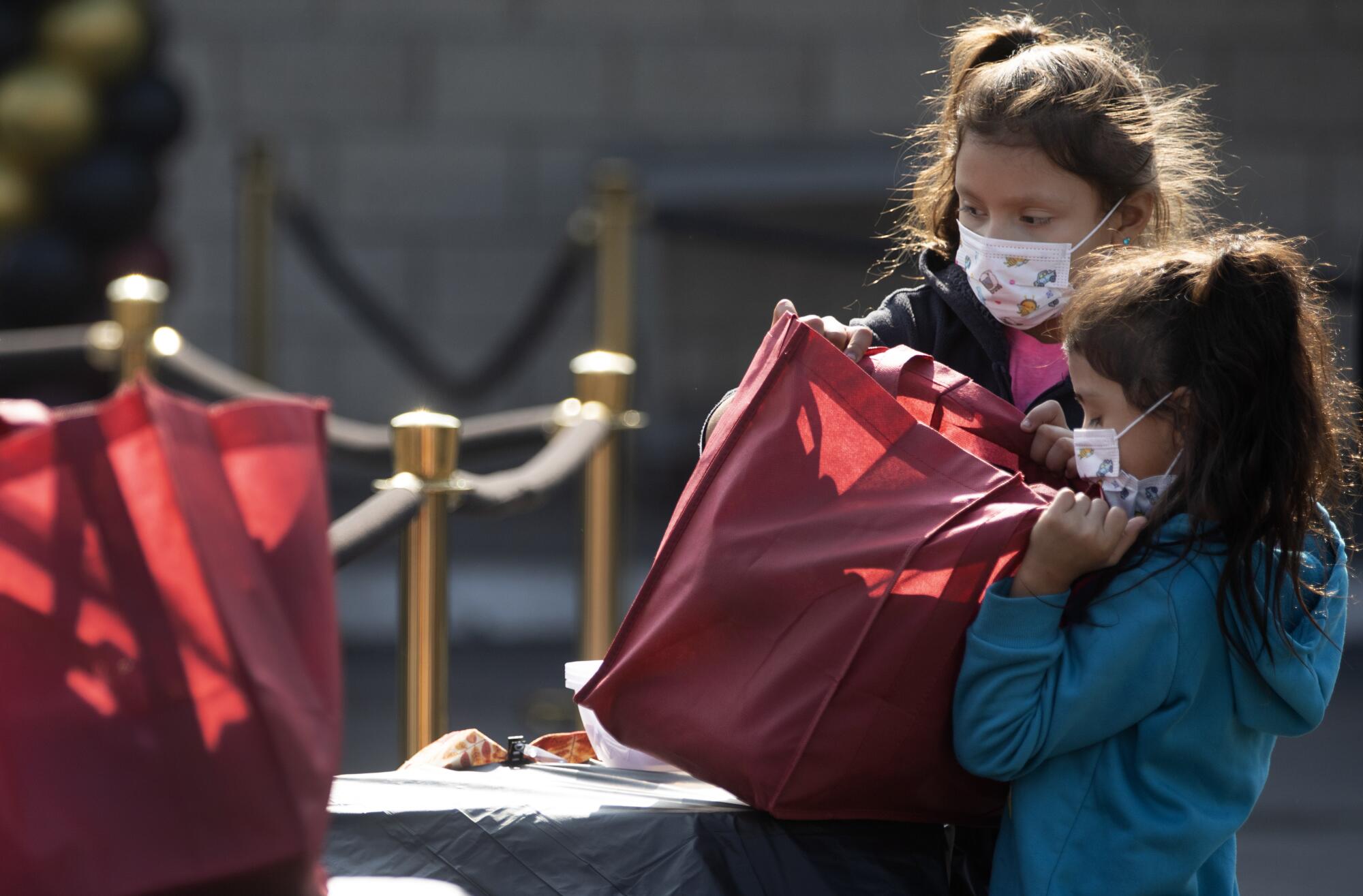 Two young girls in masks pick up a red tote bag filled with food and hygiene products