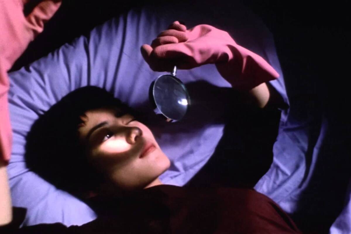 A woman lies on her back in a bed, holding a magnifying glass over her face, her hand in a pink rubber glove.