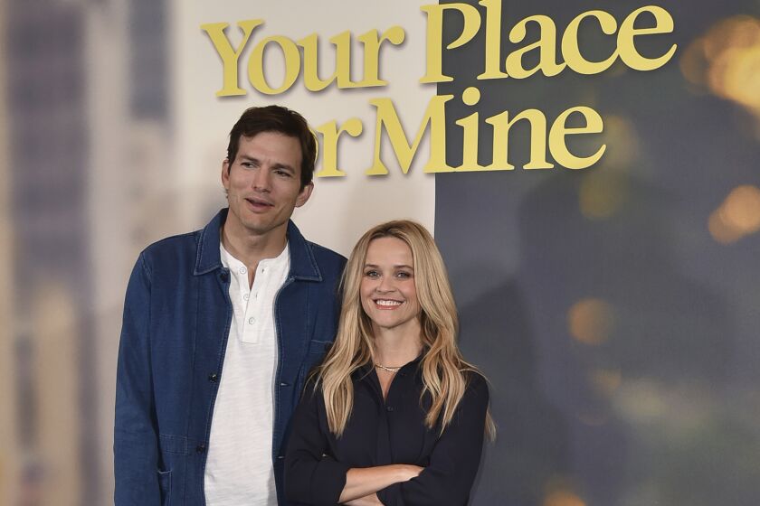 Ashton Kutcher in a blue jacket and brown pants standing next to Reese Witherspoon in a black dress