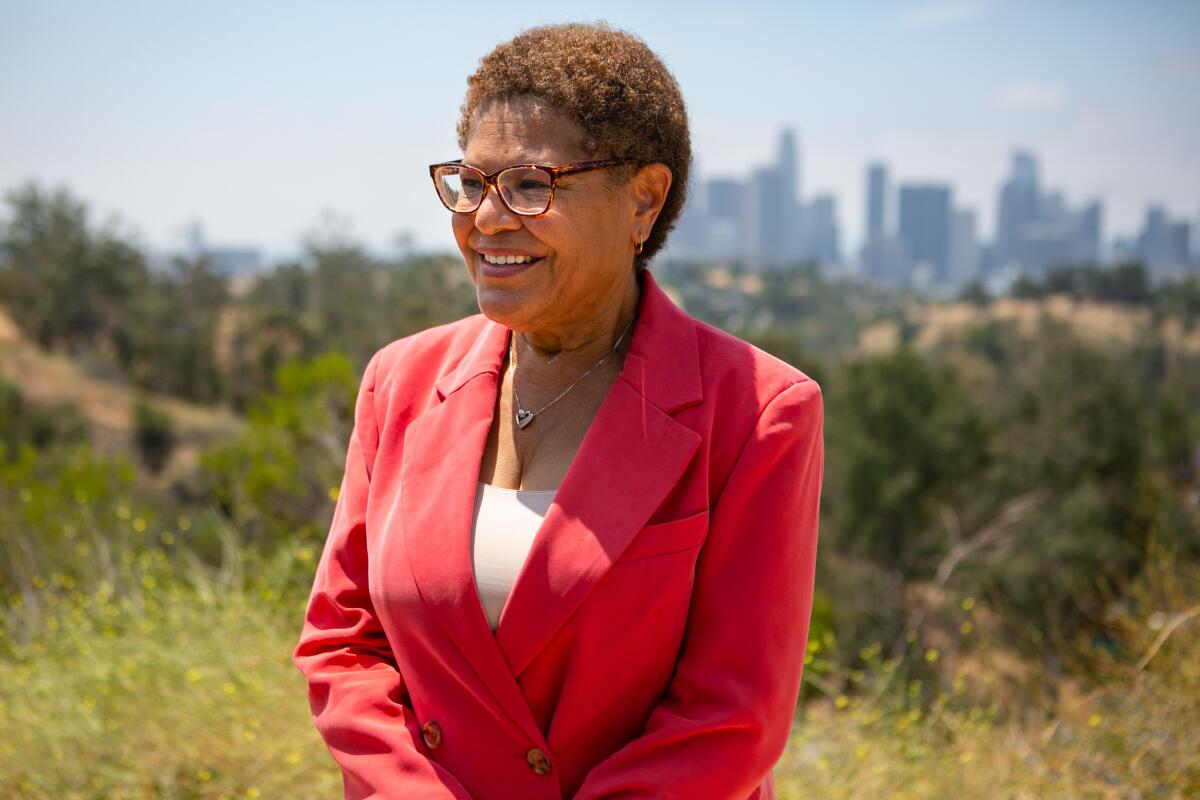 Karen Bass stands wearing a red suit with the downtown L.A. skyline in the distance.