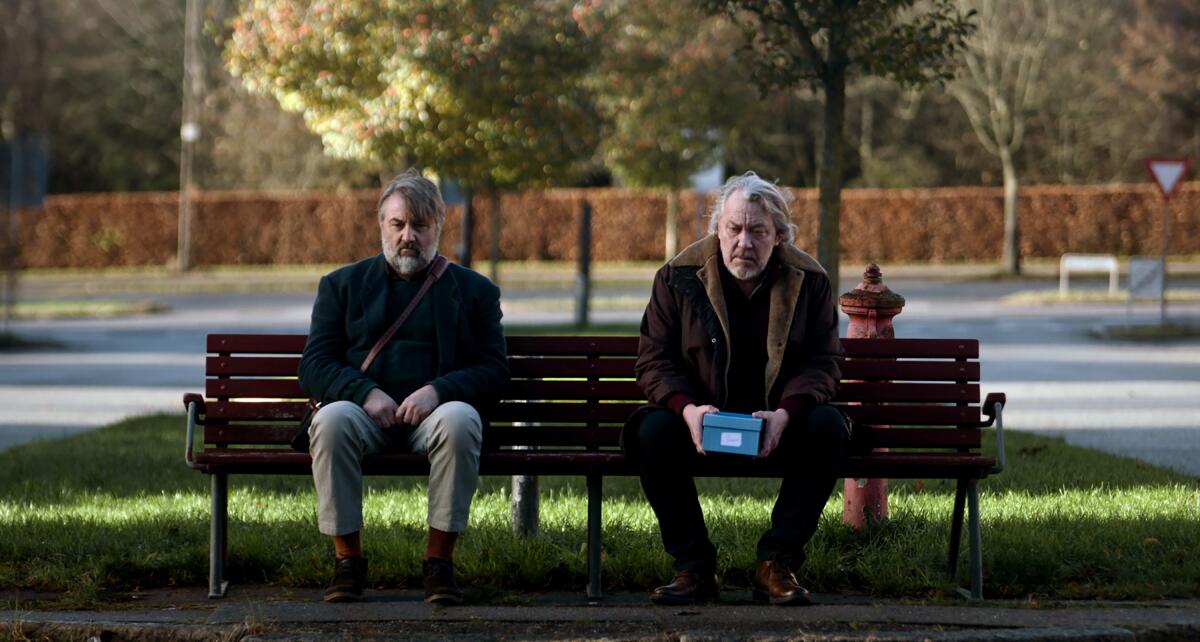 Two older men sit on a park bench, one holding a box in a scene from the short film "Knight of Fortune."