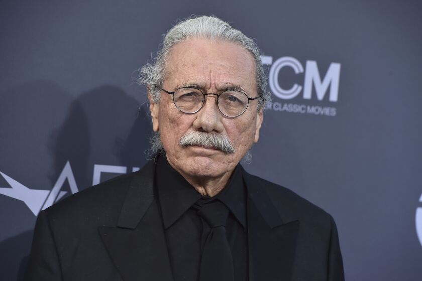 Edward James Olmos poses in a black suit and tie with round, black-rimmed glasses.