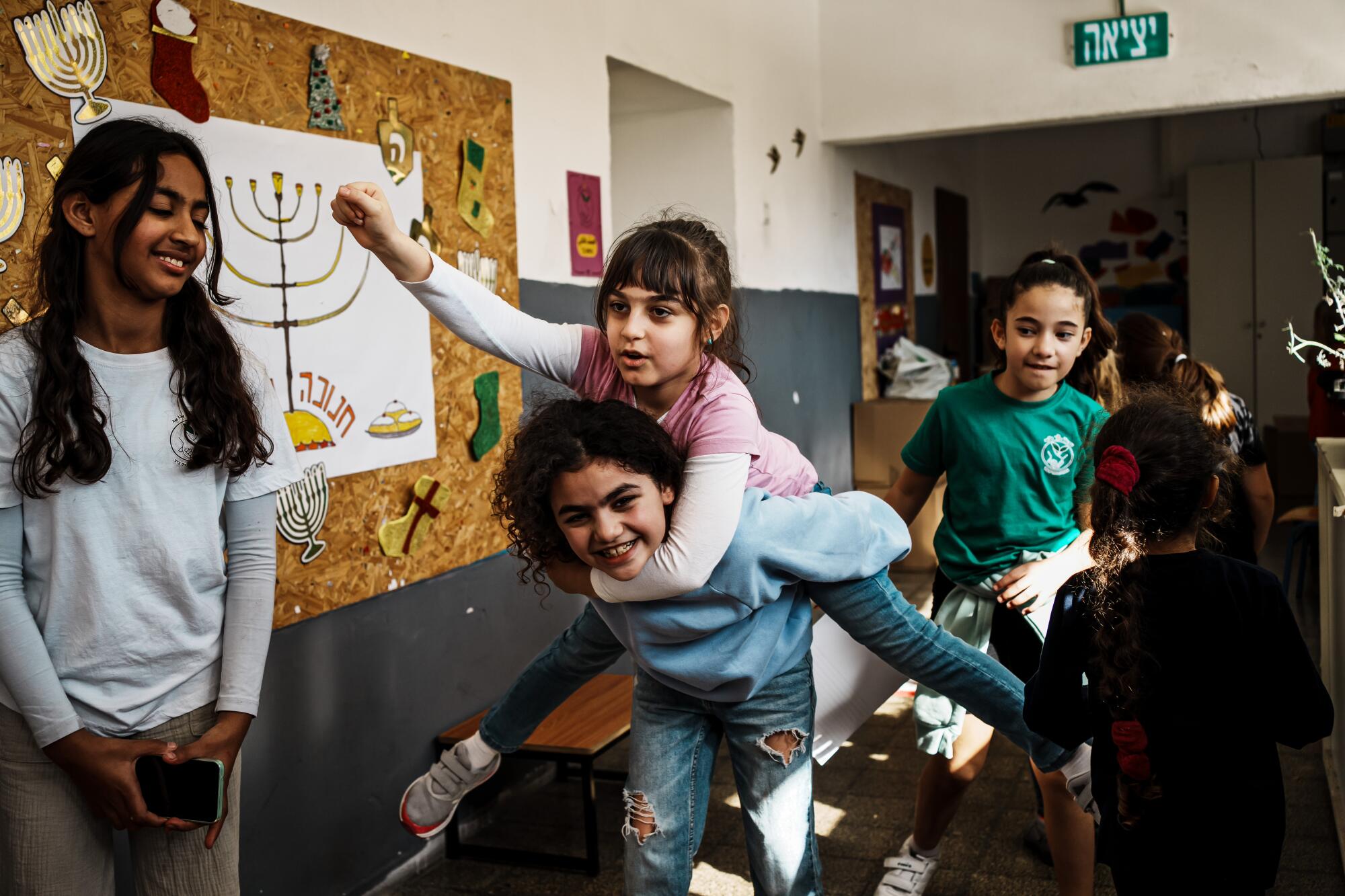 A girl carries another girl on piggyback near a drawing of a menorah on the wall as other girls watch 