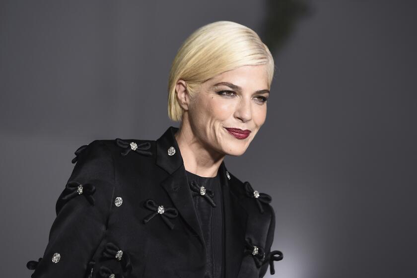 Selma Blair with short platinum hair posing in a black suit and red lipstick