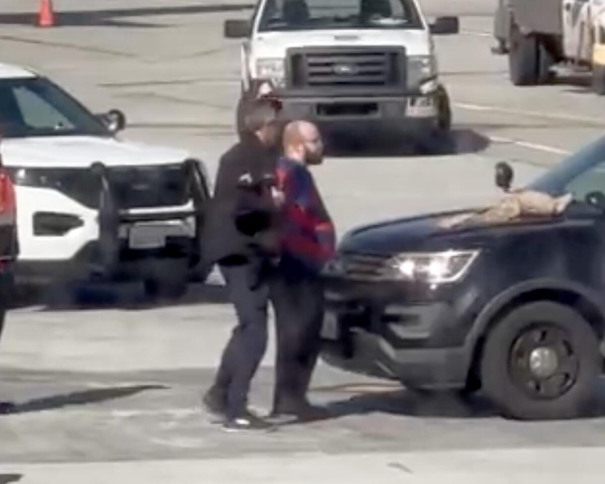 A man is standing between police cars with another man behind, holding him by his shoulders. 