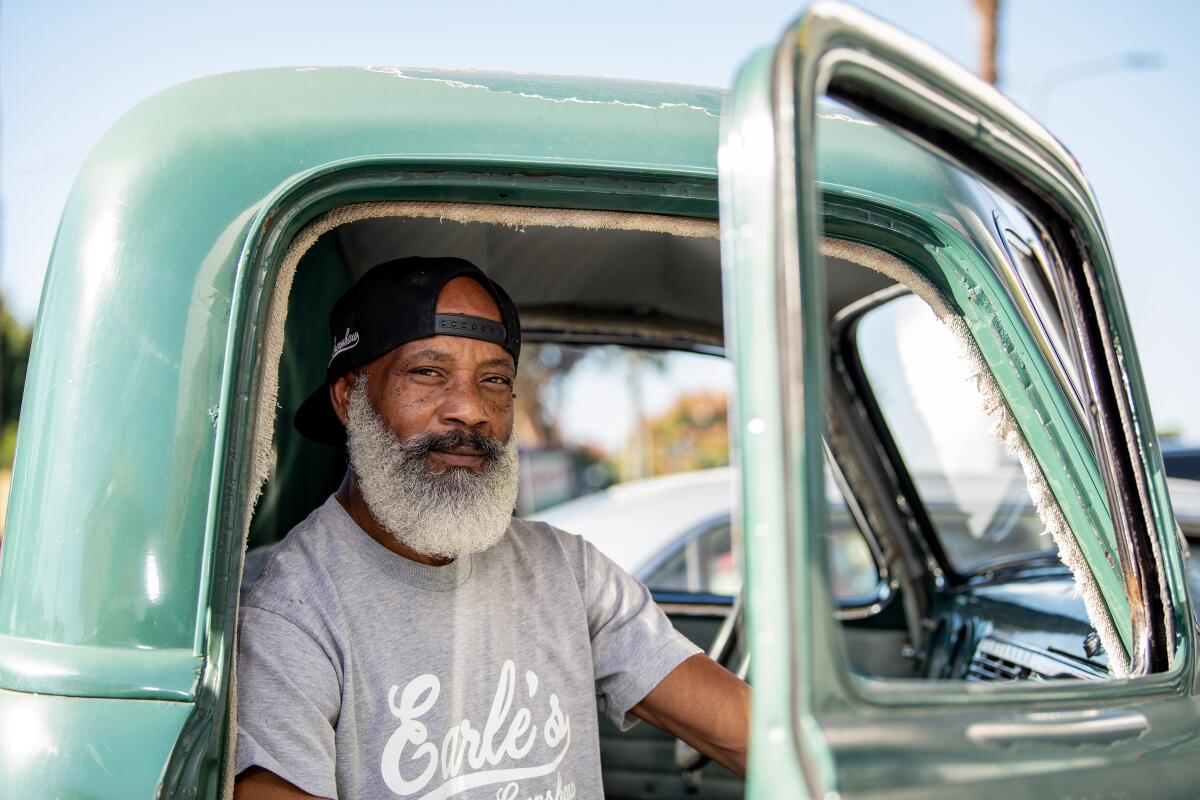 A Black man with a white beard, wearing an Earle's T-shirt, seated in an aqua pickup truck, its door ajar