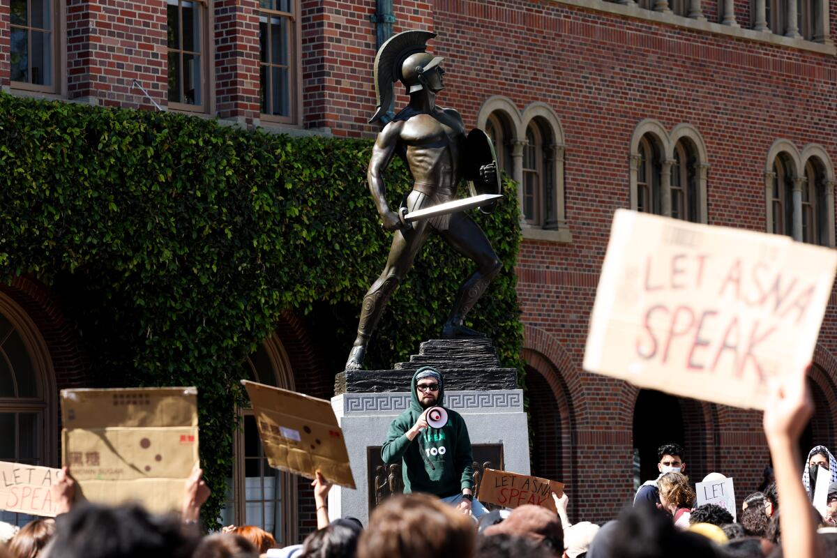 A person with a megaphone stands below a Trojan statue, facing a crowd with signs including one reading, "Let Asna speak"